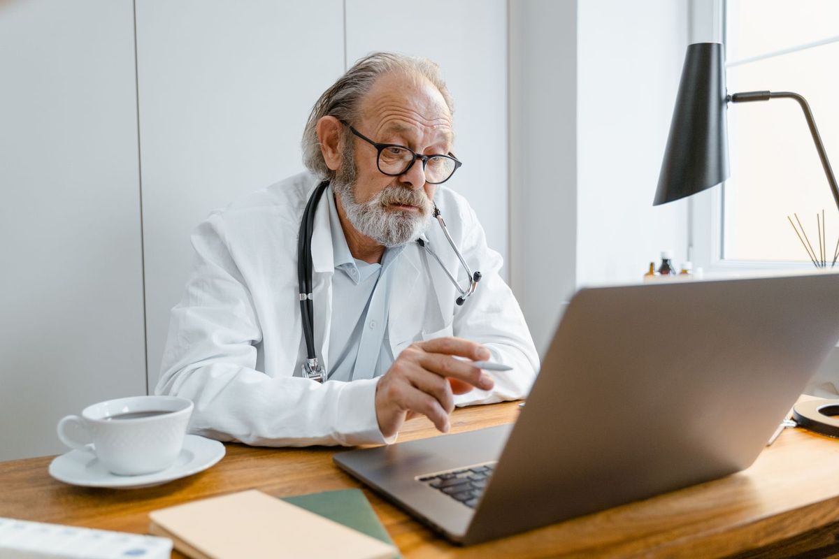 A clinician with a stethoscope reviewing patient information on a laptop at his desk