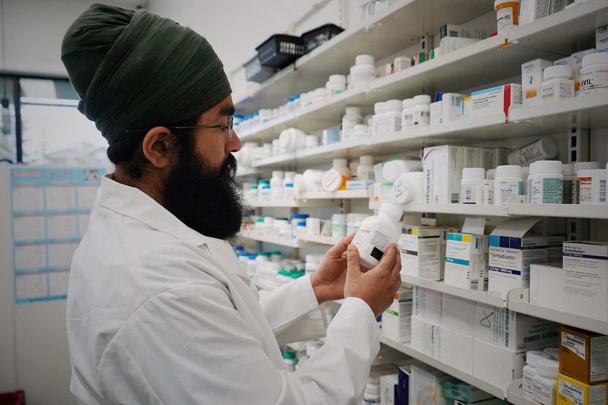 A pharmacist in a white coat checking a medication bottle in front of pharmacy shelves